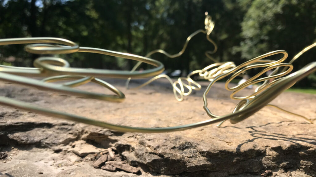 A close up of a length of a length of gold wire sitting on a natural stone surface. The wire is shaped into swirling and waved patterns. The sunlight hits the sculpture, throwing a shadow on to the stone. In the background is a green forest, out of focus.